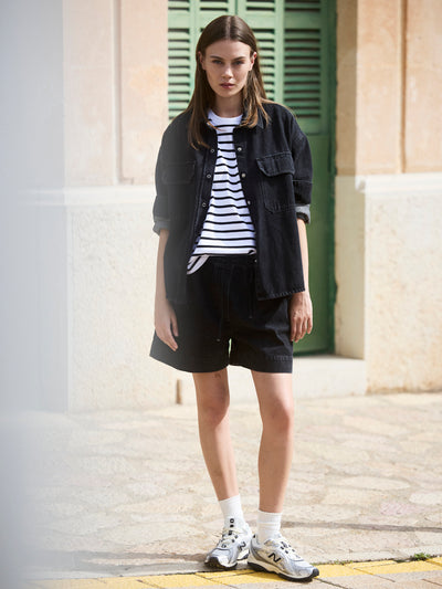 Damen Black Denim Overshirt aus Bio-Baumwolle, vorne getragen über einem schwarz-weiß gestreiften T-Shirt und Denim-Shorts.
