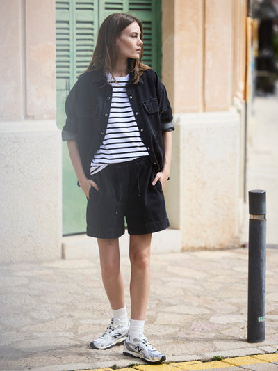 Damen Black Denim Overshirt aus Bio-Baumwolle in seitlicher Ansicht, getragen über gestreiftem T-Shirt und schwarzen Denim-Shorts.