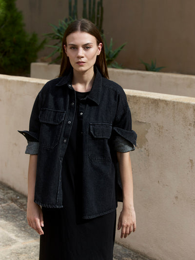 Damen Black Denim Overshirt aus Bio-Baumwolle im Porträt, offen getragen mit gerollten Ärmeln über schwarzem Kleid.