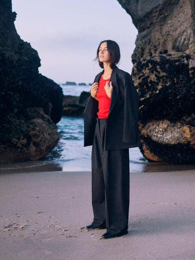 Model mit rotem Top und schwarzem WOTE Blazer aus nachhaltiger Tuchwolle, stehend am Strand zwischen Felsen, Blick seitlich.
