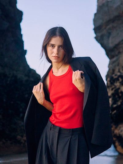 Nahaufnahme eines Models im schwarzen WOTE Blazer aus nachhaltiger Tuchwolle über rotem Top, fotografiert zwischen Felsen am Strand.