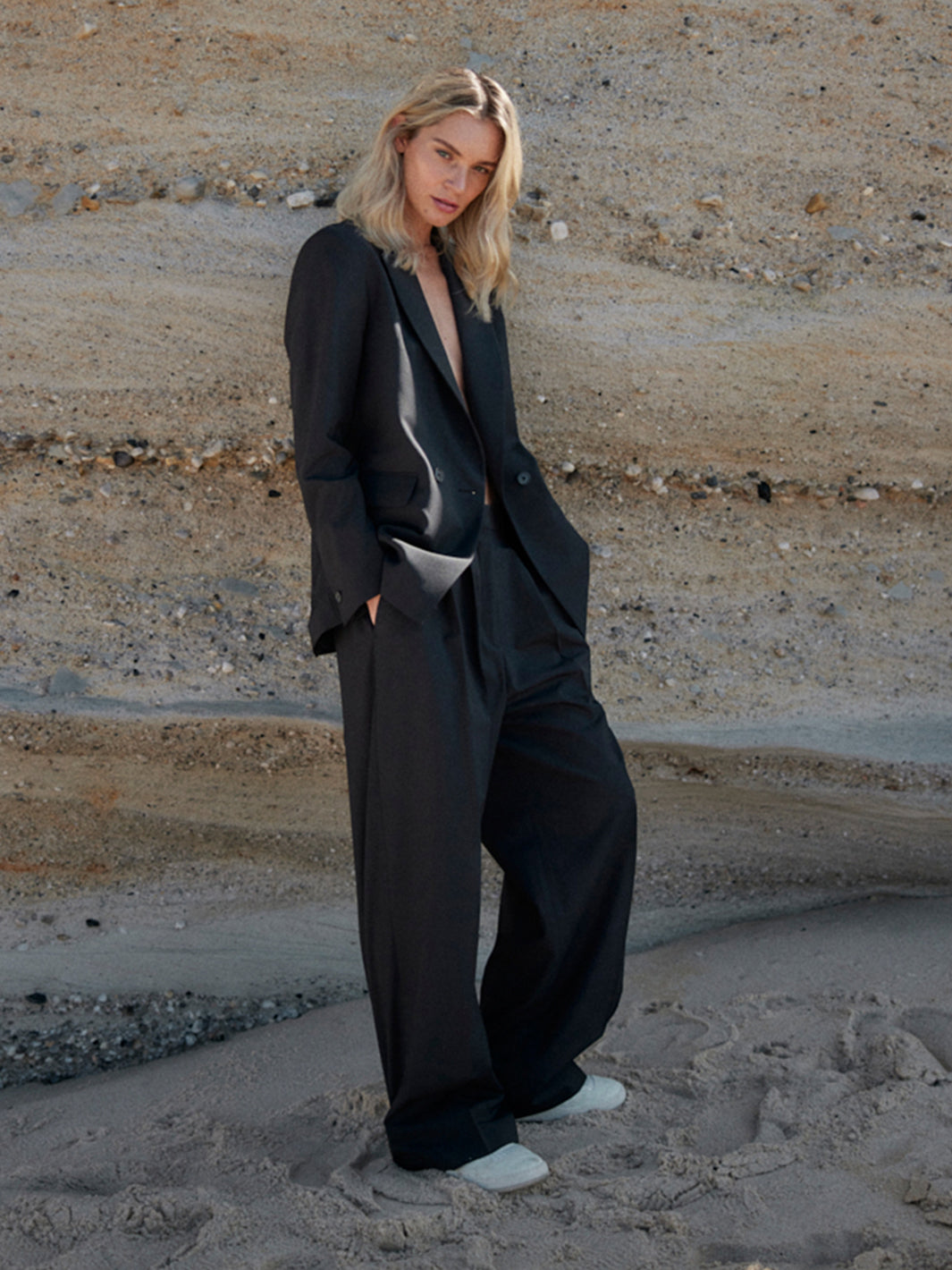 Model trägt schwarzen WOTE Blazer aus nachhaltiger Tuchwolle vor geschichteter Sandsteinwand am Strand.