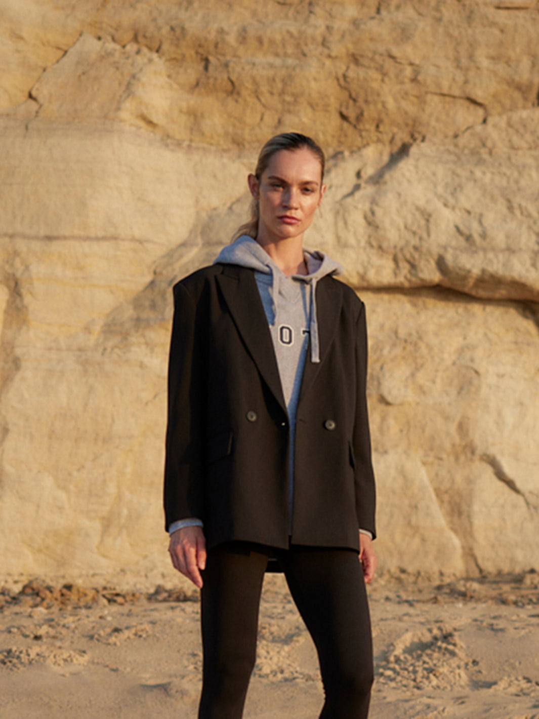 Model trägt schwarzen WOTE Blazer aus nachhaltiger Tuchwolle über grauem Hoodie, stehend vor heller Klippenwand im warmen Abendlicht.