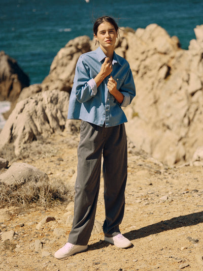 Frau trägt eine graue Classic Hose aus zertifizierter Wolle mit Bundfalten und lockerem Denimhemd vor Felsen am Meer.
