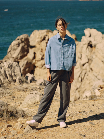 Frau trägt eine graue Classic Hose aus zertifizierter Wolle mit Bundfalten und lockerem Denimhemd, stehend vor Felsen am Meer.