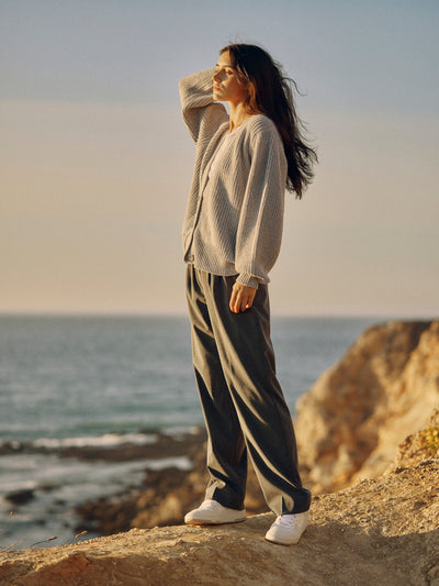 Frau trägt eine graue Classic Hose aus zertifizierter Wolle mit Bundfalten und hellen Strickcardigan, stehend an der Küste mit Meerblick.