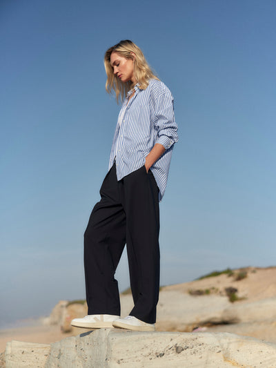 Frau trägt eine schwarze Classic Hose aus zertifizierter Wolle und ein blau-weiß gestreiftes Hemd, stehend auf einer Felsklippe am Strand.