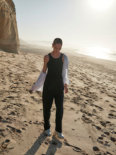 Frau trägt eine schwarze Classic Hose aus zertifizierter Wolle mit schwarzem Top und offenem weißen Hemd, beim Gehen am sonnigen Strand.