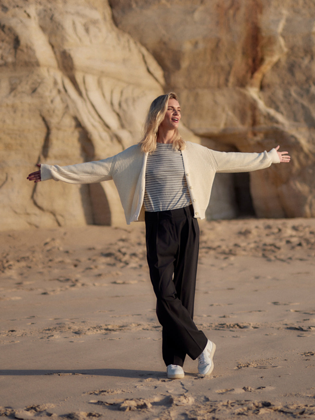 Frau trägt eine schwarze Classic Hose aus zertifizierter Wolle am Strand, locker kombiniert mit gestreiftem Shirt und heller Strickjacke.