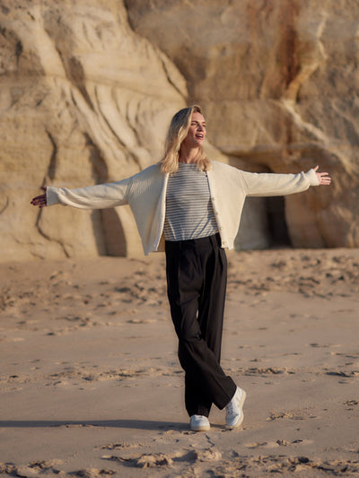 Frau trägt eine schwarze Classic Hose aus zertifizierter Wolle am Strand, locker kombiniert mit gestreiftem Shirt und heller Strickjacke.