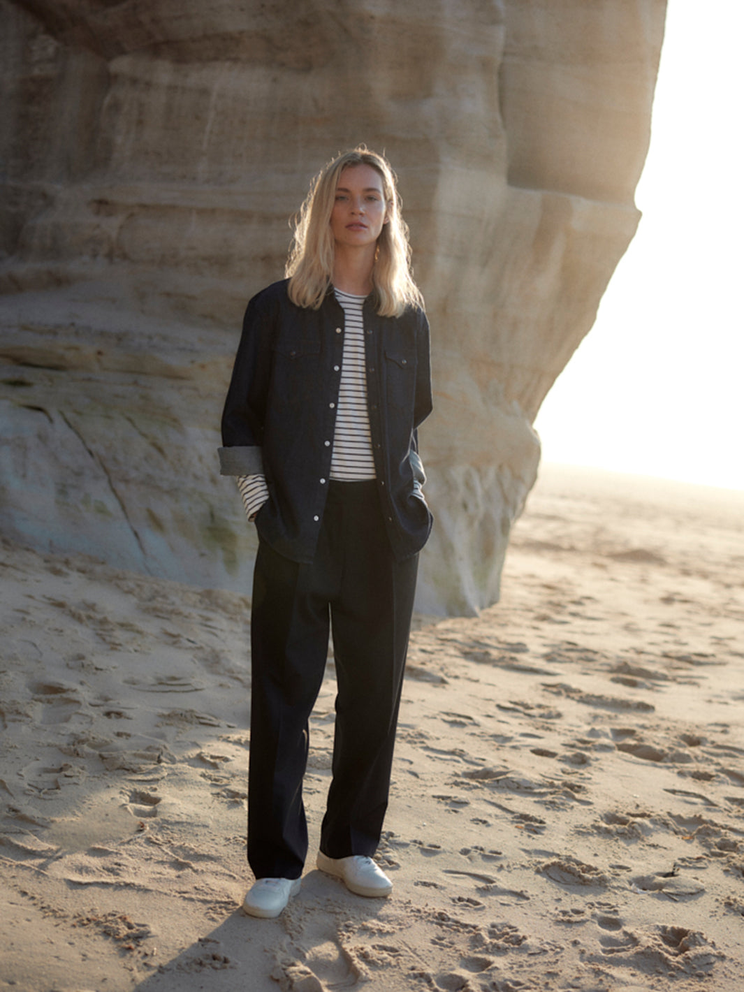 Frau trägt eine schwarze Classic Hose aus zertifizierter Wolle, kombiniert mit gestreiftem Shirt und dunkelblauem Jeanshemd, stehend am Strand vor einer Felsformation.