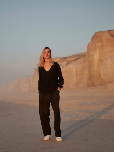 Frau trägt eine schwarze Classic Hose aus zertifizierter Wolle und einen schwarzen V-Ausschnitt-Pullover, gehend am Strand bei Sonnenuntergang.