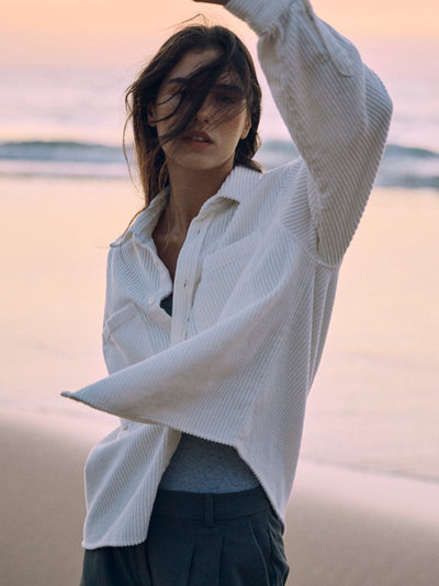 Frau trägt ein weißes Cord-Overshirt aus Organic Cotton am Strand bei Sonnenuntergang, leicht bewegtes Haar und lockerer, moderner Look.