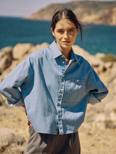 Model trägt eine kurze hellblaue Denimbluse aus Bio-Baumwolle vor einer Felsküste mit Meer im Hintergrund.