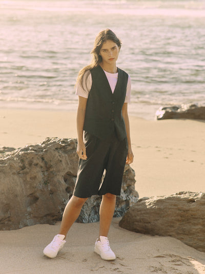 Modell trägt eine schwarze Bermuda aus zertifizierter Wolle im frontalen Look zwischen Felsen am Strand.