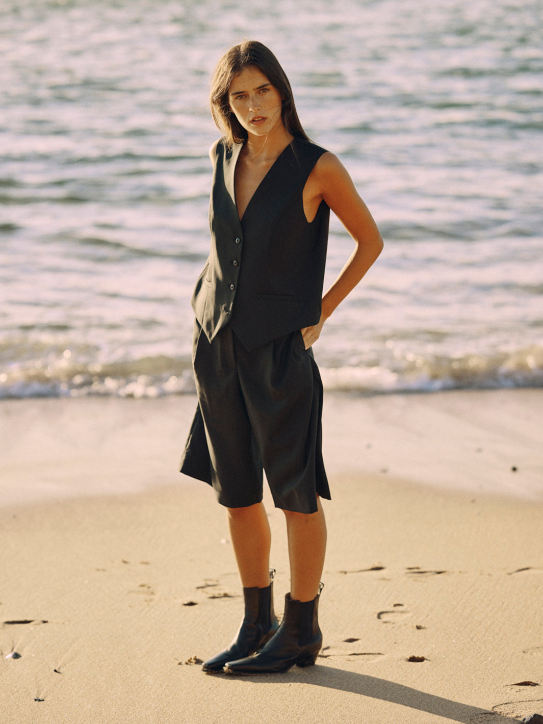 Modell trägt eine schwarze Bermuda aus zertifizierter Wolle im Ganzkörper-Look am Strand.