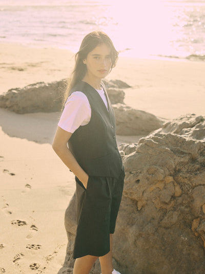 Modell trägt eine schwarze Bermuda aus zertifizierter Wolle in seitlicher Ansicht am Felsenstrand.