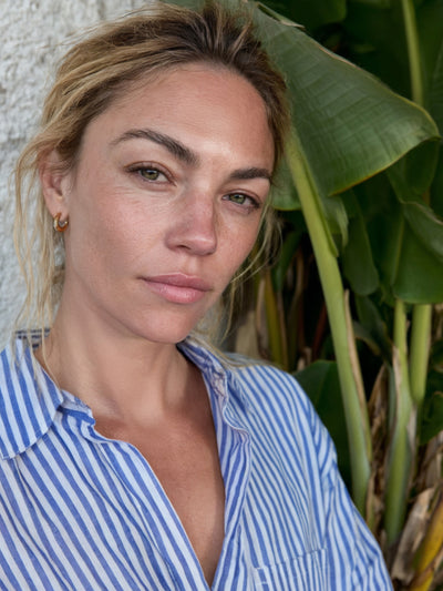 Nahaufnahme einer Frau im blau-weiß gestreiften Midi-Hemdblusenkleid aus Organic Cotton vor großen grünen Blättern.