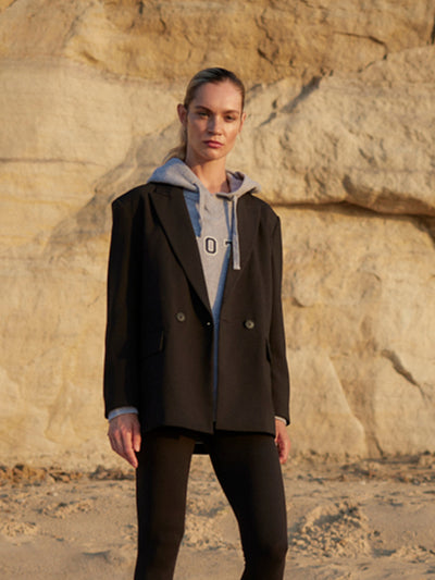 Model trägt einen grauen WOTE Logo Hoodie aus Organic Cotton unter einem schwarzen Blazer, stehend vor heller Felskulisse.