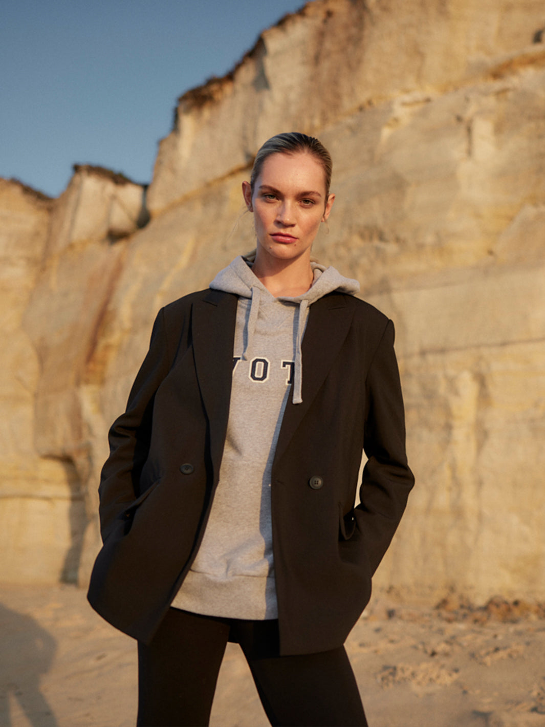 Model trägt einen grauen WOTE Logo Hoodie aus Organic Cotton unter einem schwarzen Blazer im Abendlicht.