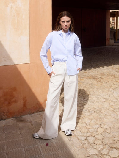 Model trägt eine weiße Wide-Leg-Hose und ein hellblaues Hemd vor einer Terracotta-Wand in mediterraner Umgebung.