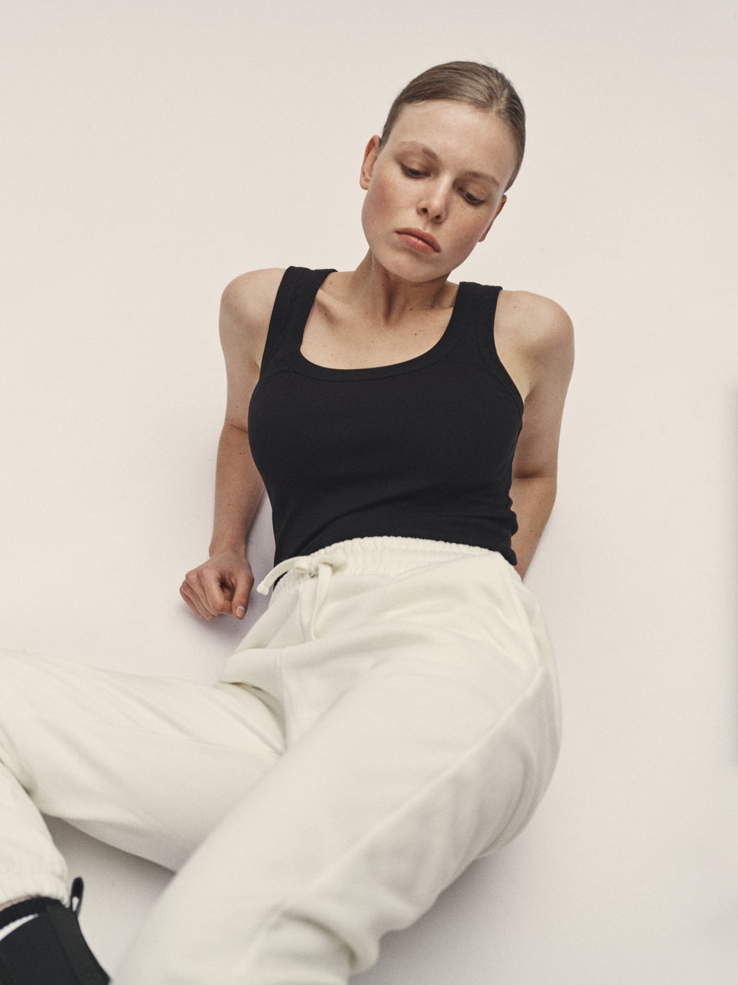 Damen Tank Top in Schwarz aus 96% Bio-Baumwolle mit 4% Elasthan, körpernaher Schnitt und tiefer Rundhalsausschnitt, nachhaltig in Portugal produziert - WOTE Classic Fit, Model im Studio mit Sweatpants in Off-White liegend